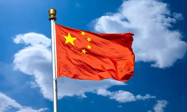 The national flag of China, featuring a red field with five yellow stars, waving against a blue sky dotted with clouds.