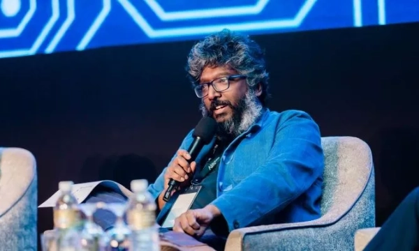 A speaker with curly hair and glasses holds a microphone while seated on a panel, discussing topics with a blue backdrop and water bottles on the table.