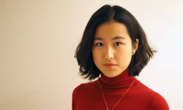 A young woman with short, wavy black hair, wearing a red turtleneck, poses in front of a plain background with a serious expression.