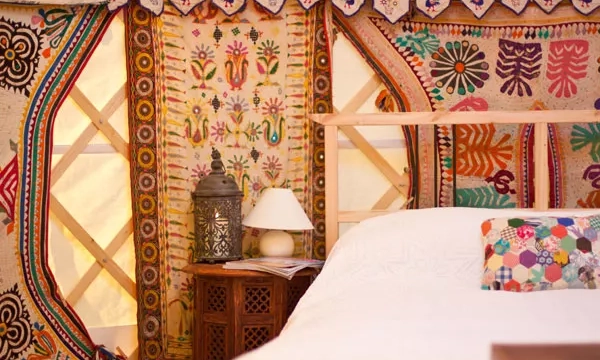 A cozy interior of a yurt featuring a bed, colorful patterned wall hangings, a wooden side table with a lamp, and decorative cushions.