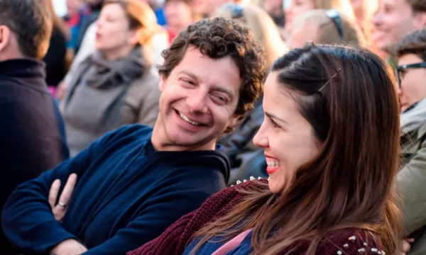 A smiling couple enjoying a lively outdoor event, surrounded by a diverse audience in the background, with warm lighting creating a cheerful atmosphere.