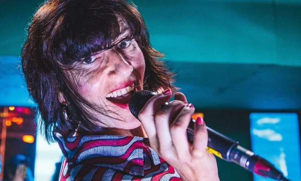 A close-up of a woman with short dark hair, holding a microphone and smiling energetically while performing on stage. Bright colors and lights in the background enhance the lively atmosphere.
