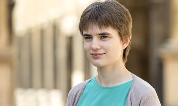 A young person with short brown hair, wearing a teal shirt and a light cardigan, standing outdoors with a blurred background of stone architecture.