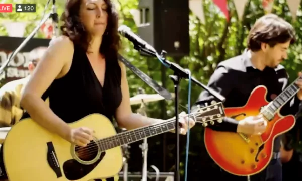A woman playing an acoustic guitar sings into a microphone, while a man in a black shirt plays an electric guitar in a lively outdoor setting.