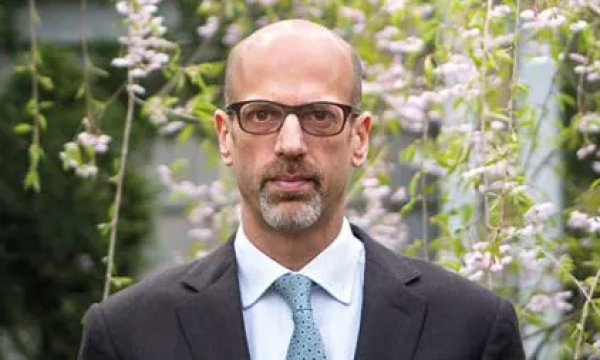 A man with glasses and a suit stands outdoors surrounded by flowering plants, with a serious expression on his face.