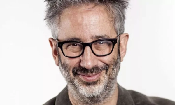 A smiling man with glasses and short, spiky gray hair, dressed in a dark jacket, against a light background.