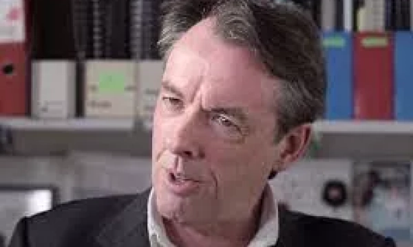 A man in a suit speaking intently, with shelves of files and folders in the background.