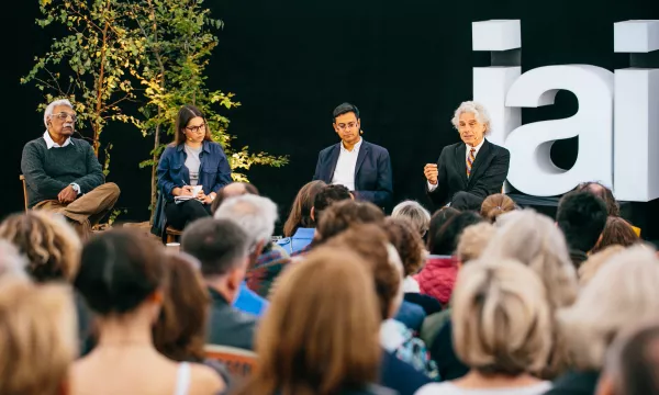 A panel discussion featuring four speakers, seated in front of an audience, with lush greenery and large letters spelling "iai" in the background.