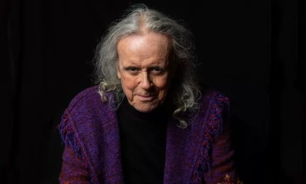 An elderly man with long, wavy gray hair, wearing a purple shawl, is smiling against a black background.
