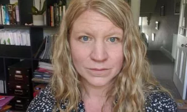 A woman with curly blonde hair and blue eyes is sitting in a room with books and papers in the background.