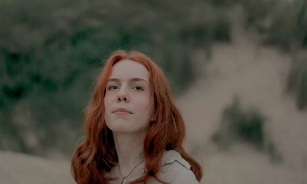 A woman with wavy red hair stands against a blurred natural background, looking confidently at the camera.
