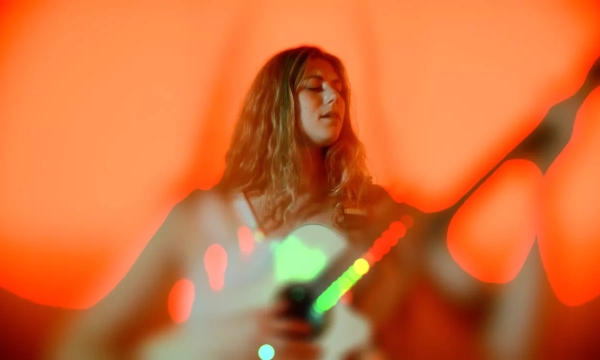 A musician with wavy hair plays a guitar in front of an orange backdrop, creating a vibrant, dreamlike atmosphere with blurred lights.