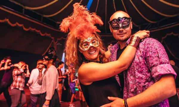 Two people wearing vibrant masks and costumes dance in a festive atmosphere, surrounded by others in a colorful, lively setting.