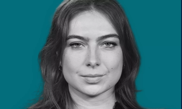 A close-up portrait of a young woman with long, dark hair and a neutral expression, set against a teal background.