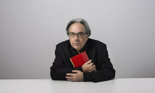 A man in a black blazer holds a red book close to his chest, seated at a white table against a plain gray background.