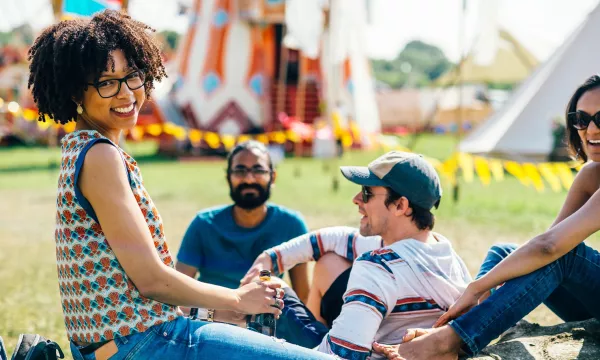 A group of four friends enjoying a sunny day outdoors, sitting on grass near colorful tents, smiling and relaxing together.