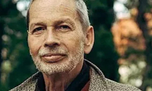 A middle-aged man with gray hair and a beard, dressed in a textured coat, stands outdoors against a backdrop of trees.
