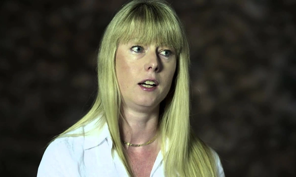 A woman with long blonde hair, wearing a white shirt, speaking thoughtfully in a softly lit setting.