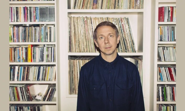 A person stands in front of shelves filled with CDs and records, wearing a dark blue shirt, looking directly at the camera.