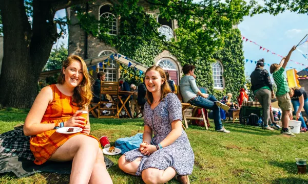 Two young women sit on the grass enjoying food and drinks at an outdoor event near a historic building, with colorful bunting and people in the background.