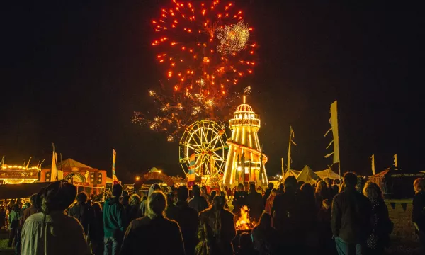 A crowd watches a vibrant fireworks display over a fairground, featuring a large Ferris wheel and a lit-up amusement ride at night.