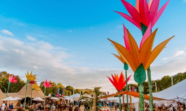 A vibrant outdoor festival scene featuring large, colorful flower sculptures, tents, and people enjoying the atmosphere under a bright blue sky.