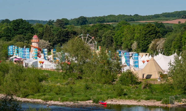 A vibrant festival scene featuring colorful tents, a ferris wheel, and greenery along a river, set against a backdrop of hills and blue skies.