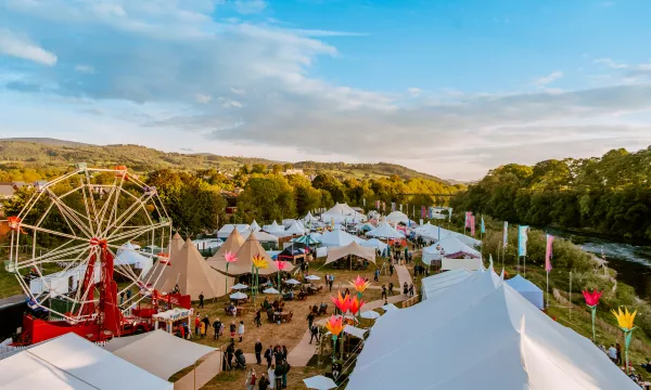 A vibrant festival scene featuring a large ferris wheel, colorful tents, and attendees enjoying various activities amidst lush greenery and a serene river backdrop.