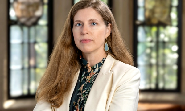 A woman with long, wavy hair and blue earrings, wearing a white blazer over a floral blouse, poses confidently in a well-lit room with large windows.
