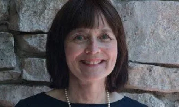 A woman with shoulder-length brown hair smiles warmly in front of a rustic stone wall. She wears a black top and a pearl necklace.