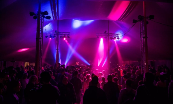 A vibrant crowd dances under colorful lights inside a large tent during a lively music event, surrounded by illuminated stage equipment.