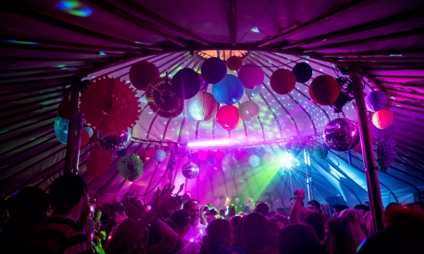 A vibrant party scene inside a tent, illuminated by colorful lights and decorated with paper lanterns, as a lively crowd dances and enjoys the atmosphere.