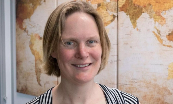 A woman with short blonde hair smiles in front of a world map mural, wearing a black and white striped top.