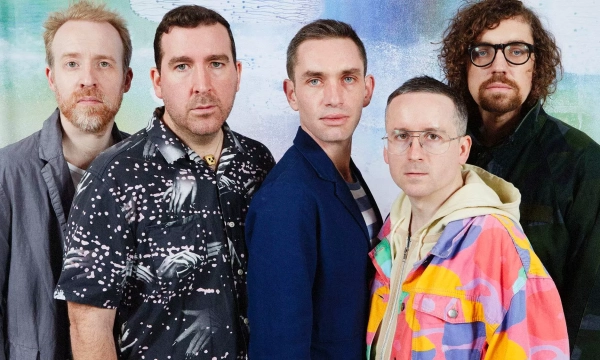 A group of five men posing together, wearing stylish and colorful clothing against an abstract, vibrant background.