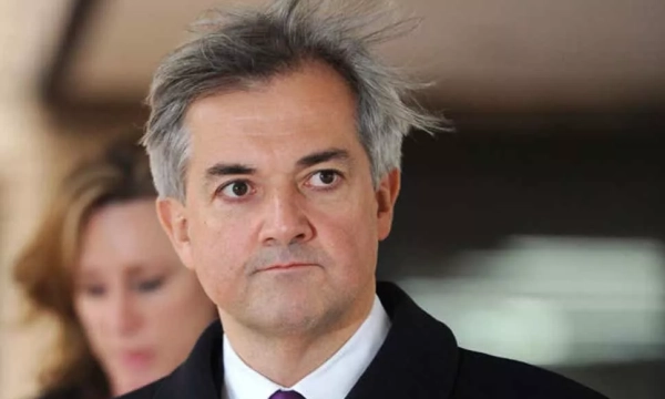 A man with disheveled gray hair wearing a dark coat and tie, looking serious in an outdoor setting, with a blurred figure in the background.