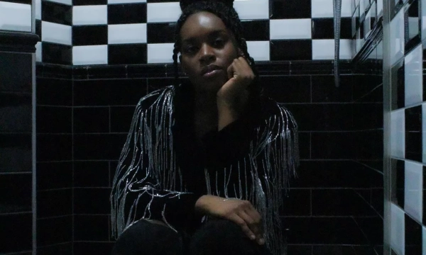 A person squats in a black and white checkered shower, wearing a fringed jacket and high-heeled boots, with a contemplative expression.
