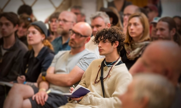 A diverse audience attentively listens during a presentation, with a young individual in a beige sweater taking notes in the foreground.