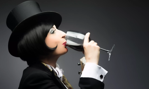 A glamorous figure in a top hat and formal attire sips wine from a glass, showcasing elegance and sophistication against a dark background.