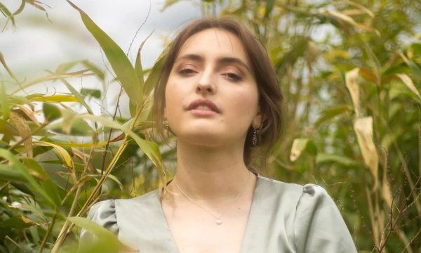 A woman in a light green dress stands among tall plants, with a serene expression against a cloudy sky backdrop.