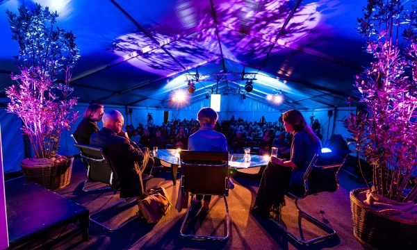 A panel discussion in a brightly lit tent with a diverse audience visible in the background, featuring four speakers seated at a table surrounded by decorative plants.