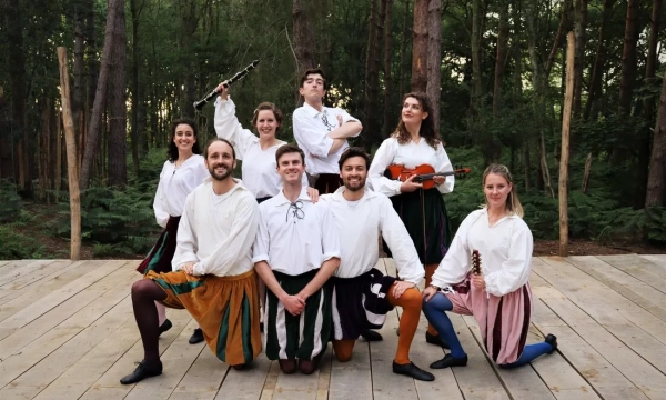 A group of nine performers dressed in Renaissance-style costumes, posing on a wooden stage in a forest, with instruments like a flute and violin.