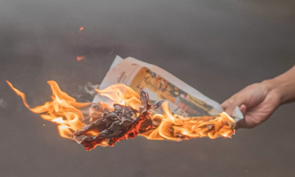 A hand holds a flaming piece of newspaper, with bright orange flames and smoke rising from it.
