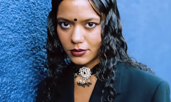 A portrait of a woman with long, wavy hair, wearing a black suit and a decorative necklace, set against a textured blue background.