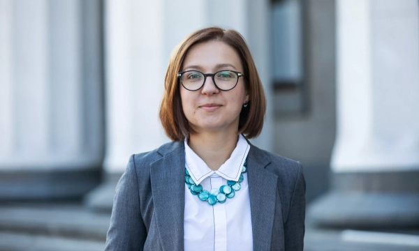 A confident woman in a gray blazer and white shirt stands outdoors, wearing a turquoise necklace and glasses, with classical architecture in the background.