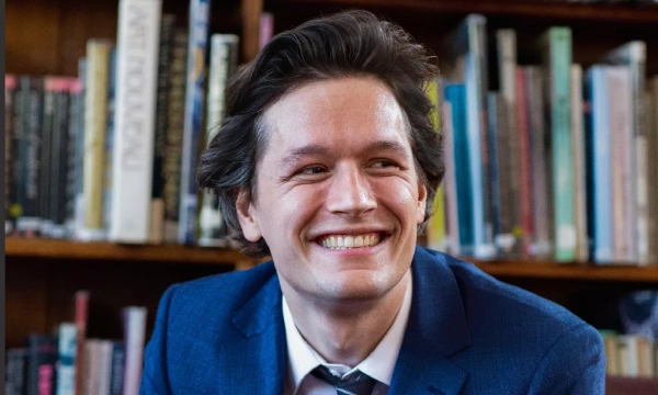 Smiling man in a blue suit, seated in front of a bookshelf filled with books.