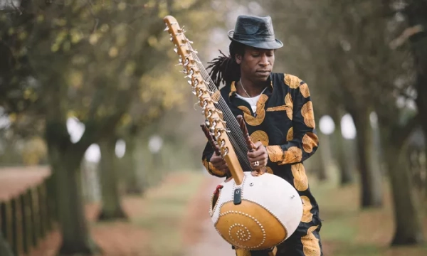A musician dressed in a patterned outfit plays a kora, a traditional West African string instrument, on a pathway lined with trees.