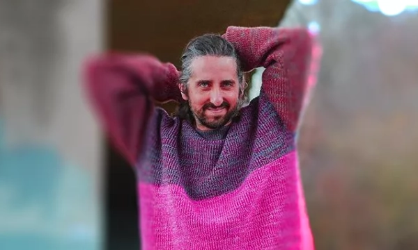 A smiling person wearing a colorful pink and purple sweater, standing outdoors with a blurred natural background.