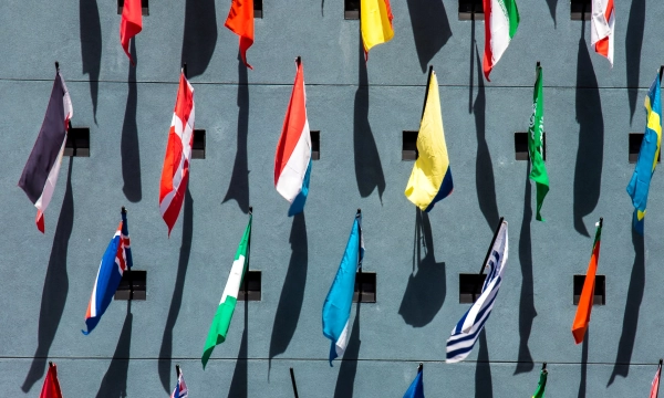 A wall displaying a variety of international flags, including Denmark, Netherlands, Italy, and Canada, casting shadows against a blue background.