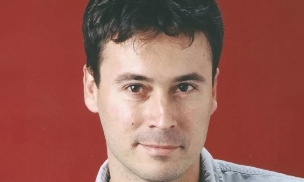 A close-up portrait of a man with dark, wavy hair and a slight smile, set against a red background.