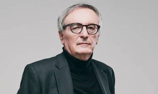 A middle-aged man with glasses and gray hair, dressed in a black turtleneck and blazer, poses against a neutral background, conveying a serious demeanor.
